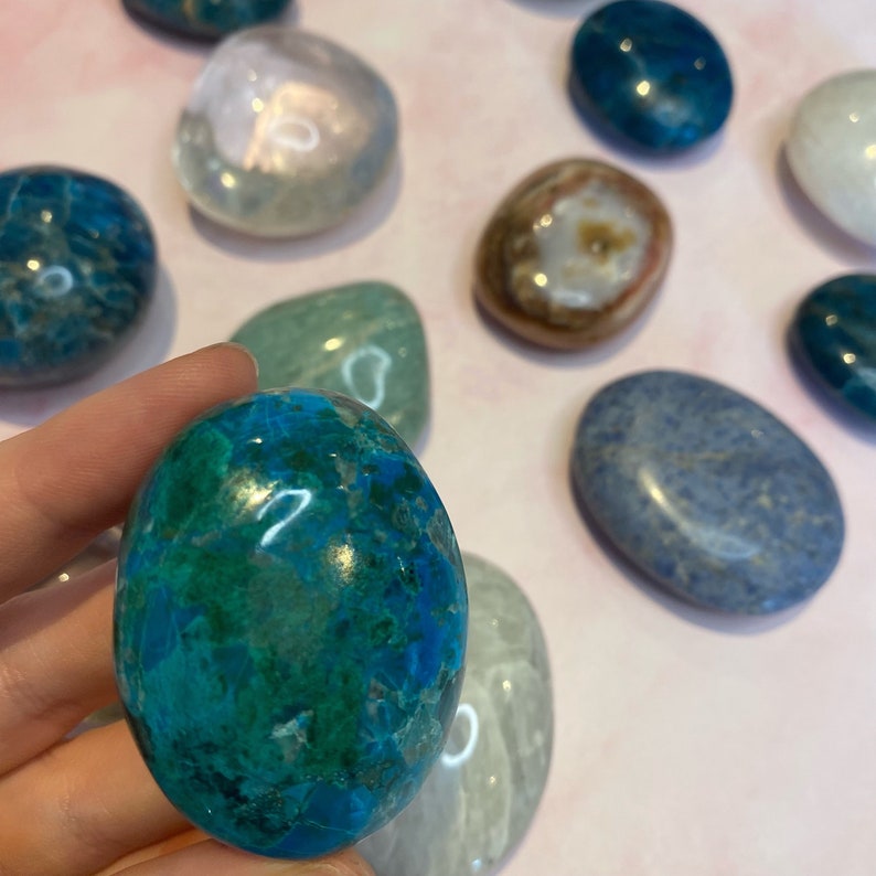 Op de afbeelding: Een verzameling gepolijste edelstenen in verschillende kleuren, waaronder blauw, groen, wit en bruin. De ovaalvormige stenen hebben een glad, reflecterend oppervlak. Een blauwgroene steen wordt vastgehouden.