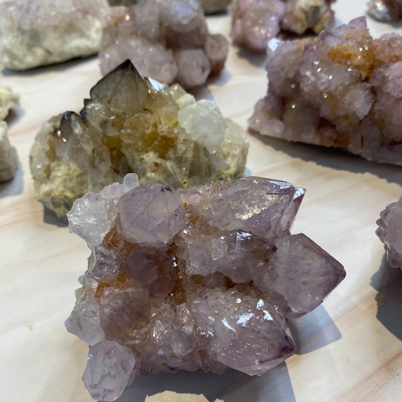 Op de afbeelding: Een verzameling amethistkristalclusters in verschillende maten en vormen. De kristallen vertonen een reeks kleuren, van licht lavendel tot diep paars, met enkele gebieden van heldere kwarts. De kristallen hebben een natuurlijke, ruwe textuur.