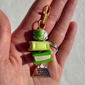 Könnte beinhalten: Ein goldfarbener Schlüsselanhänger mit einem Anhänger mit einem vierblättrigen Kleeblatt, zwei Miniaturbüchern und einem bergförmigen Anhänger. Der Anhänger trägt den Text "Faith can move mountains". Die Bücher sind grün und gelb.