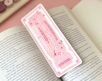 May include: A pink and white bookmark with a ticket design. It says "Lifetime Member of the Cowboy Romance Book Club" and "Boots & Hat Required". It also says "Admit One Cowgirl".
