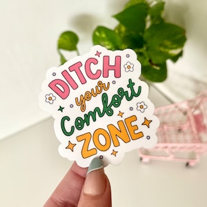 May include: A pink, green, and yellow sticker with the text "Ditch your comfort zone" in a whimsical font. The sticker features small white flowers and stars.