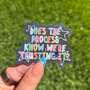 May include: A black die-cut sticker featuring the colorful text "DOES THE PROCESS KNOW WE'RE TRUSTING IT?" surrounded by white outlined star shapes. The sticker has a dark background with small colorful speckles and a holographic finish.