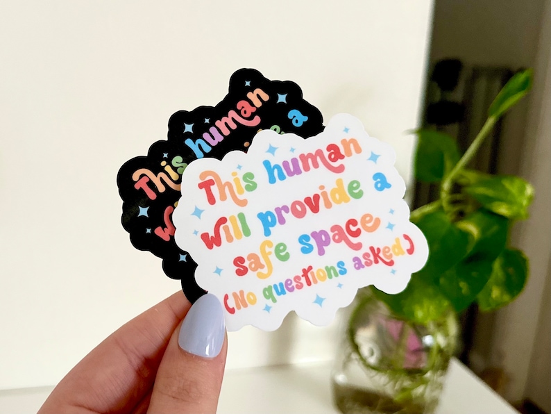 Op de afbeelding: Een witte sticker met regenboogkleurige tekst die "This human will provide a safe space (No questions asked)" luidt. De sticker heeft de vorm van een wolk met sterren rond de tekst.