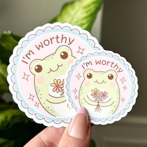 May include: Two pink and blue circular stickers with a white scalloped border. Each sticker features a green frog holding a bouquet of flowers and the text "I'm worthy".