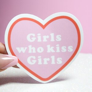 Puede incluir: Pegatina rosa en forma de coraz&oacute;n con un contorno blanco y un borde rojo. La pegatina dice "Girls who kiss Girls" en texto blanco.