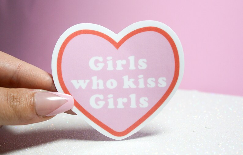 Puede incluir: Pegatina rosa en forma de coraz&oacute;n con un contorno blanco y un borde rojo. La pegatina dice "Girls who kiss Girls" en texto blanco.