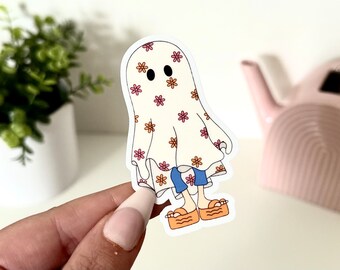 Puede incluir: Una pegatina de fantasma blanco con un patrón floral y pantalones vaqueros azules. El fantasma lleva zapatos naranja y blancos.