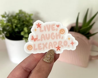 Pode incluir: Adesivo rosa e laranja com o texto "Live, Laugh, Lexapro." O adesivo tem um design de flor, borboleta e rosto sorridente.