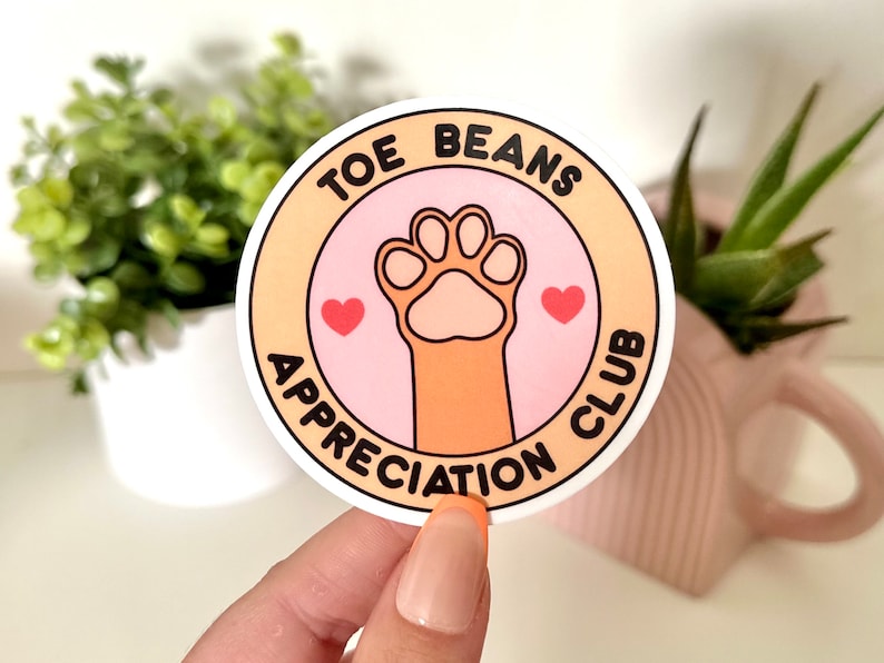 Pu&ograve; includere: Un adesivo rosa e bianco con il testo "Toe Beans Appreciation Club" e un'impronta di zampa di cartone animato con due cuori rossi.