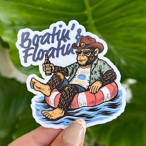 May include: A sticker featuring a cartoon monkey wearing a cowboy hat and sunglasses, holding a beer and floating on a red and white inner tube in blue water. The text "Boat'n & Floatin'" is above the monkey.