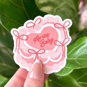 May include: Pink heart-shaped sticker with white lace trim and red bows. The sticker says "Aries Baby" in red cursive lettering.