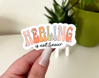 May include: A white sticker with the text "Healing is not linear" in black script. The word "Healing" is in rainbow colors with white daisy flowers around the letters.