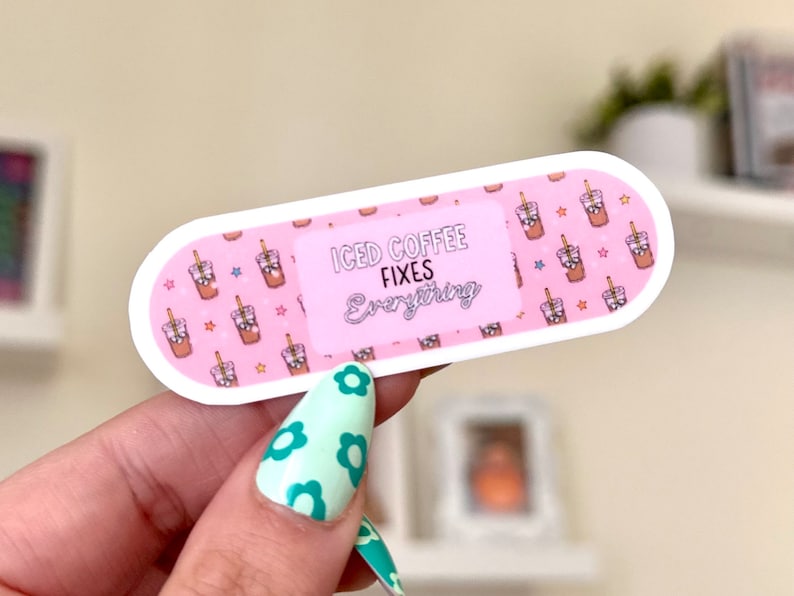 Pode incluir: Adesivo de bandagem rosa e branco com um desenho de caf&eacute; gelado de desenho animado e o texto "Iced Coffee Fixes Everything".