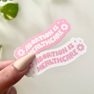 May include: Two pink and white stickers with the text "Abortion is Healthcare" in a wavy font. Each sticker has a small white flower with a yellow center.