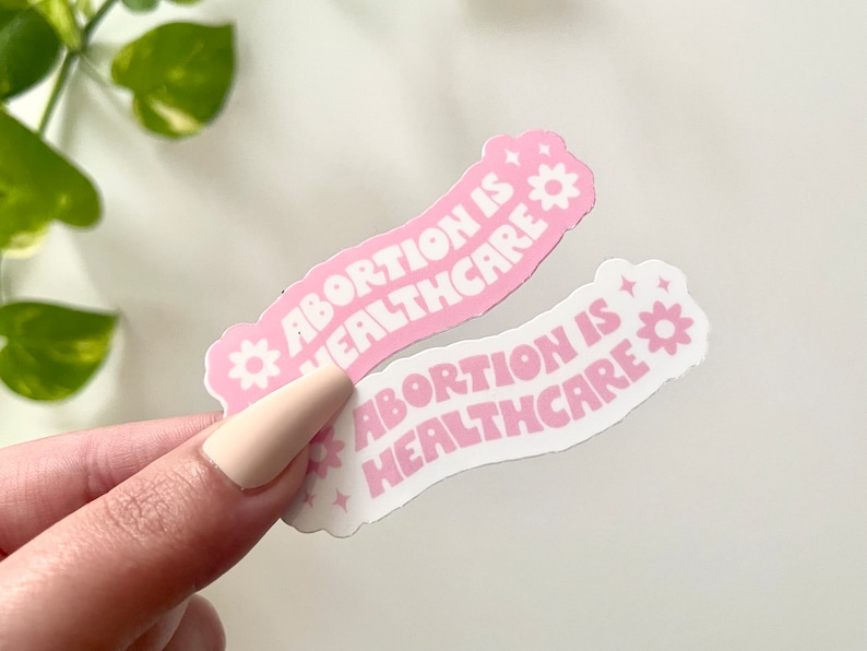 May include: Two pink and white stickers with the text "Abortion is Healthcare" in a wavy font. Each sticker has a small white flower with a yellow center.