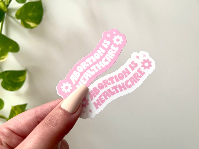 May include: Two pink and white stickers with the text "Abortion is Healthcare" in a curved font. The stickers have a pink background with white text and a white background with pink text.