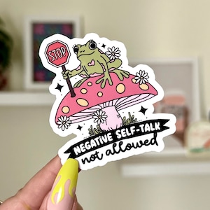 May include: A sticker with a green frog sitting on a pink mushroom holding a red stop sign. The sticker has the text "NEGATIVE SELF-TALK not allowed" in black.