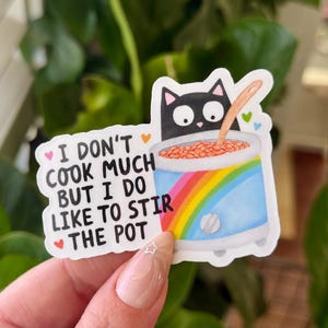 May include: A sticker featuring a black cat peering over a blue pot with a rainbow design. The pot contains a brown substance and a wooden spoon. Text on the sticker reads, "I don't cook much but I do like to stir the pot."