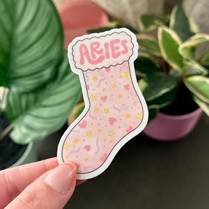 May include: Pink stocking with a white fluffy cuff and the word "Aries" in pink letters. The stocking is decorated with pink hearts, yellow flowers, and purple swirls.
