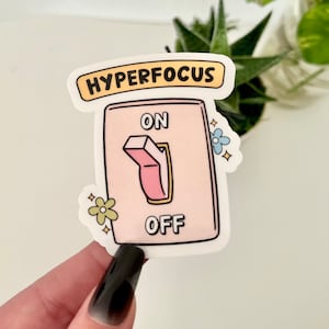 Op de afbeelding: Een roze en witte sticker met een lichtschakelaarontwerp. De schakelaar is gemarkeerd met "ON" en "OFF" en heeft een roze schuifknop. De sticker zegt "HYPERFOCUS" in gele letters.
