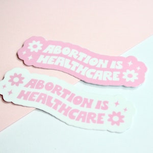 May include: Two pink and white stickers with the text "Abortion is Healthcare" in white lettering. The stickers have a flower and star design.