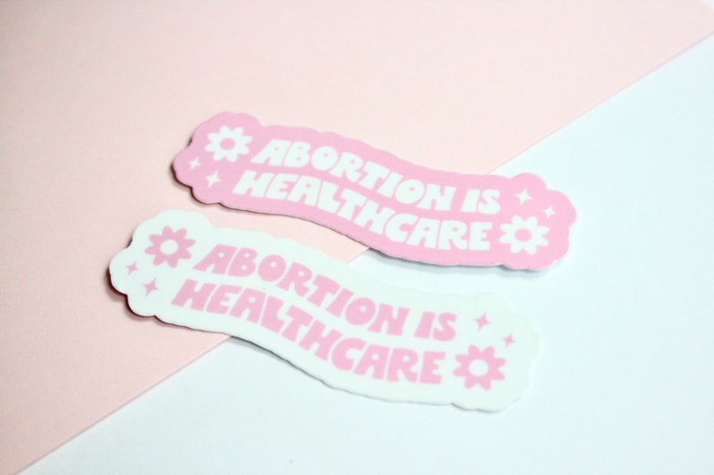 May include: Two pink and white stickers with the text "Abortion is Healthcare" in white lettering. The stickers have a flower and star design.