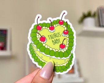 May include: A green and white heart-shaped cake with the text "TAURUS BABY" on top. The cake is decorated with red cherries and green leaves.