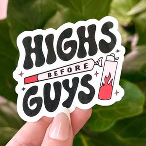 Op de afbeelding: Een witte sticker met de tekst "HIGHS BEFORE GUYS" in een zwarte retro-lettertype. De sticker heeft een afbeelding van een aangestoken sigaret en een aansteker met rode vlammen. De sticker wordt door een hand vastgehouden.