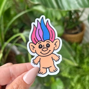 May include: A sticker of a cartoon troll with orange skin, large ears, and a smiling face. The troll's hair is pink, purple, and blue. The sticker is set against a blurred background of green plants.