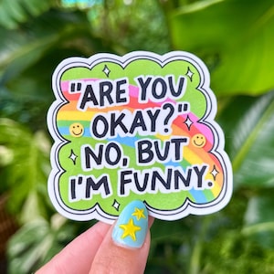 May include: A colorful sticker with the text "ARE YOU OKAY?" and "NO, BUT I'M FUNNY." The sticker has a green cloud-like border, a rainbow background, and small star and smiley face details. The sticker is held up by a person.