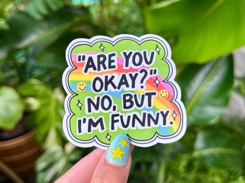 May include: A colorful sticker with the text "ARE YOU OKAY?" and "NO, BUT I'M FUNNY." The sticker has a green cloud-like border, a rainbow background, and small star and smiley face details. The sticker is held up by a person.