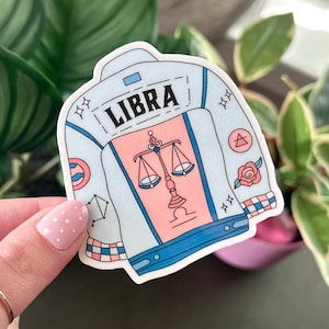 May include: A blue and pink sticker with a denim jacket design. The jacket has the word "LIBRA" on the back and a scale symbol on the front. The sticker has a white background and is decorated with stars, a rose, and a triangle.