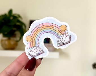 May include: A rainbow sticker with a book flying out of it. The rainbow is iridescent and has a glitter effect. There are two smiley faces on either side of the rainbow. The book is open and has a white background.