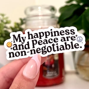 May include: A white sticker with the text "My happiness and peace are non-negotiable." The sticker has a smiley face and peace sign graphic. The sticker is held by a person with a pink and white manicure.