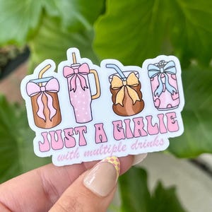 May include: A sticker featuring four stylized drink containers with bows and straws. The sticker reads "JUST A GIRLIE with multiple drinks" in pink and white bubble letters. The drinks are brown, pink, and patterned.