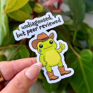 Può includere: Un adesivo con un ranocchio dei cartoni animati con cappello e stivali da cowboy, che fa il pollice in su. Il ranocchio è verde chiaro con la pancia gialla e una lacrima. Il testo "undiagnosed, but peer reviewed" è in bianco.