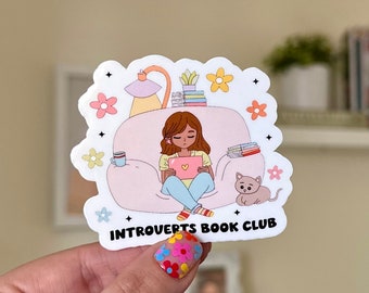 Può includere: Un adesivo bianco con un'illustrazione di un cartone animato di una persona seduta su una sedia che legge un libro. L'adesivo dice "Introverts Book Club".