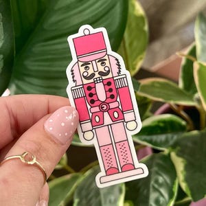 Puede incluir: Una pegatina rosa y blanca de un soldado cascanueces. El cascanueces tiene un sombrero, uniforme y botas rosas, con bigote y cejas negras. La pegatina se sostiene frente a un fondo de plantas verdes.