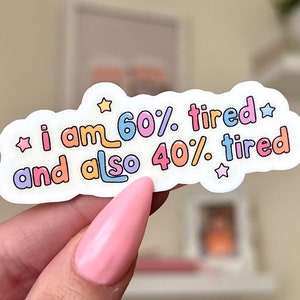 May include: A white sticker with the text "i am 60% tired and also 40% tired" in colorful bubble letters. The sticker has a cloud-like shape with star accents. The sticker is held by a person with pink nail polish.