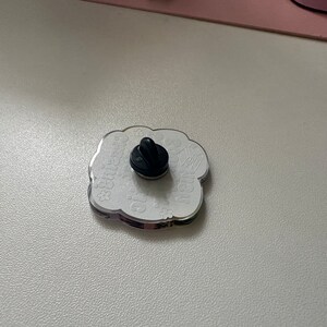 以下が含まれることがあります： 黒いピン留め具が付いた白いエナメルピン。ピンは雲の形をしていて、「Greetings from」と「The Moon」の文字が書かれています。