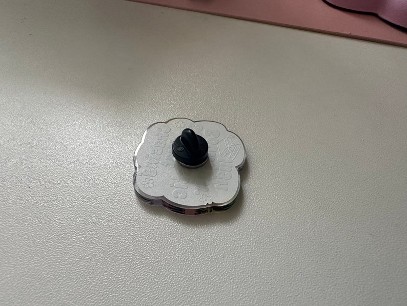 以下が含まれることがあります： 黒いピン留め具が付いた白いエナメルピン。ピンは雲の形をしていて、「Greetings from」と「The Moon」の文字が書かれています。