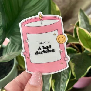 May include: A pink candle sticker with a white label that says "Smells Like A Bad Decision". The candle has a yellow smiley face on the side and a small flame on top.