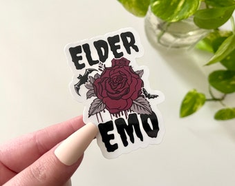 Puede incluir: Una pegatina en blanco y negro con el texto "Elder Emo" en una fuente gótica. Una rosa roja con sangre goteando y un murciélago se presentan en el diseño.