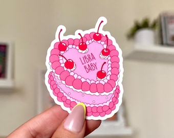 May include: Pink heart-shaped cake with white frosting and red cherries. The cake has the text "LIBRA BABY" written on it.