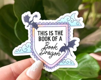 May include: A sticker with a white background and a purple, pink, and blue border. The sticker features a shield with the text "This is the book of a Book Dragon" and two black dragons flying around the shield. The sticker is surrounded by clouds.