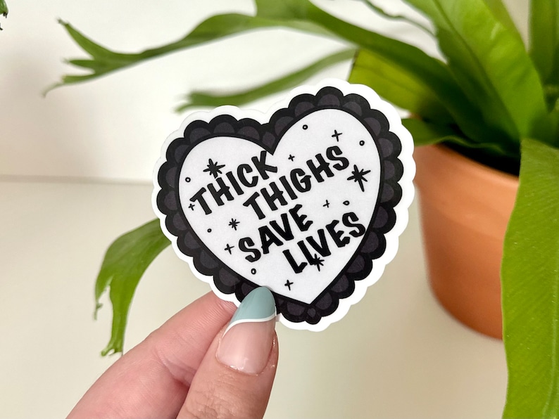 May include: A black and white heart-shaped sticker with the text "Thick Thighs Save Lives" in a bold, stylized font. The sticker has a scalloped edge and is decorated with small stars and plus signs.