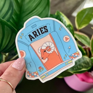 May include: A blue denim jacket sticker with a cartoon ram wearing a red and pink shirt. The jacket has a patch that says "Aries" in black text.