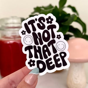 May include: White sticker with black text that reads "It's not that deep" in a retro font. The text is surrounded by small black flowers and smiley faces.