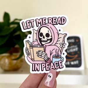 May include: A sticker featuring a cartoon skeleton wearing a pink hoodie and reading a book. The skeleton is holding a cup of coffee and is surrounded by stars. The text on the sticker reads "Let me read in peace."