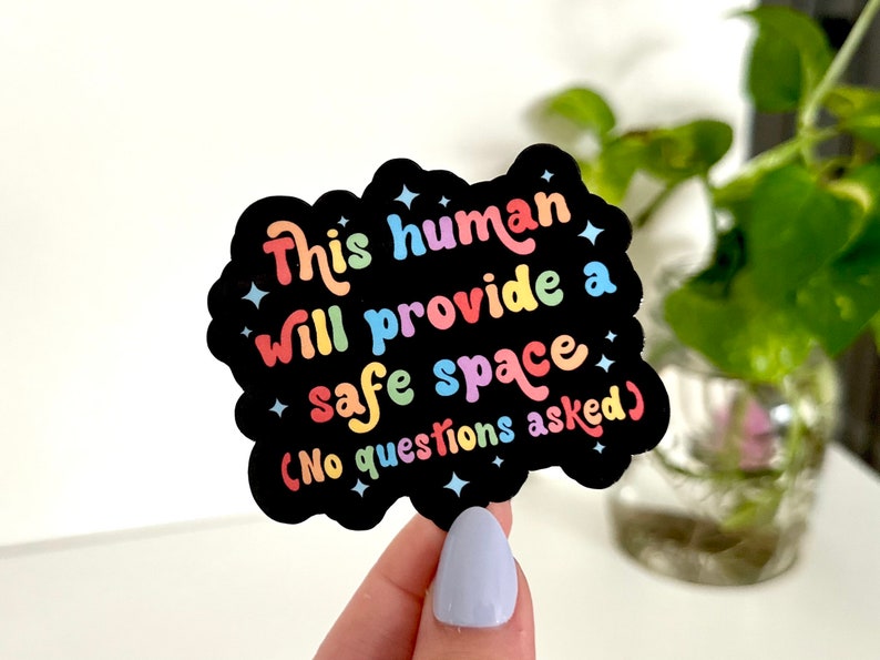 Op de afbeelding: Een zwarte sticker met regenboogkleurige tekst die "This human will provide a safe space (No questions asked)" zegt.